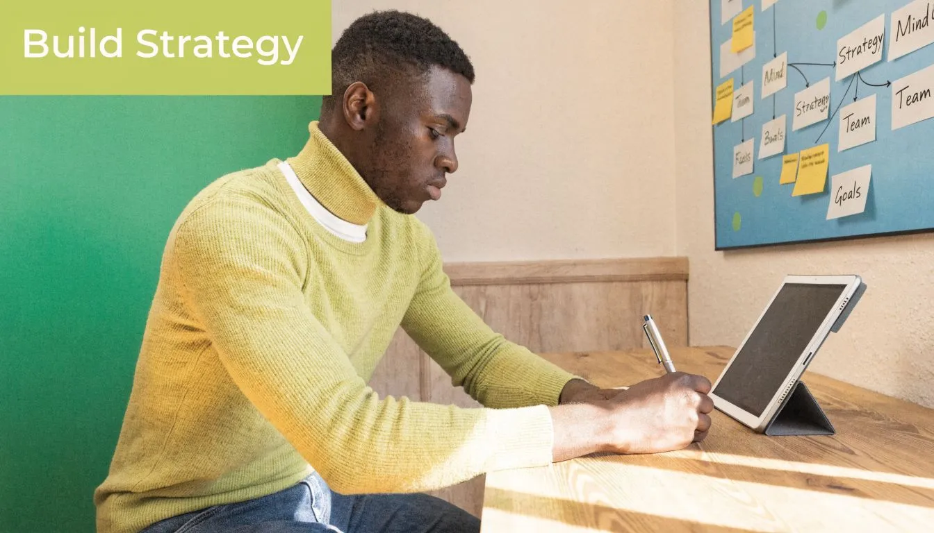 A young man sitting at a desk while writing in a notebook next to his tablet.